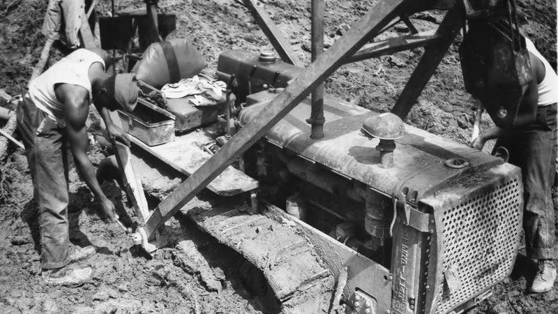 A bulldozer sunk deep in mud on the Alaska Highway, with soldiers working around it